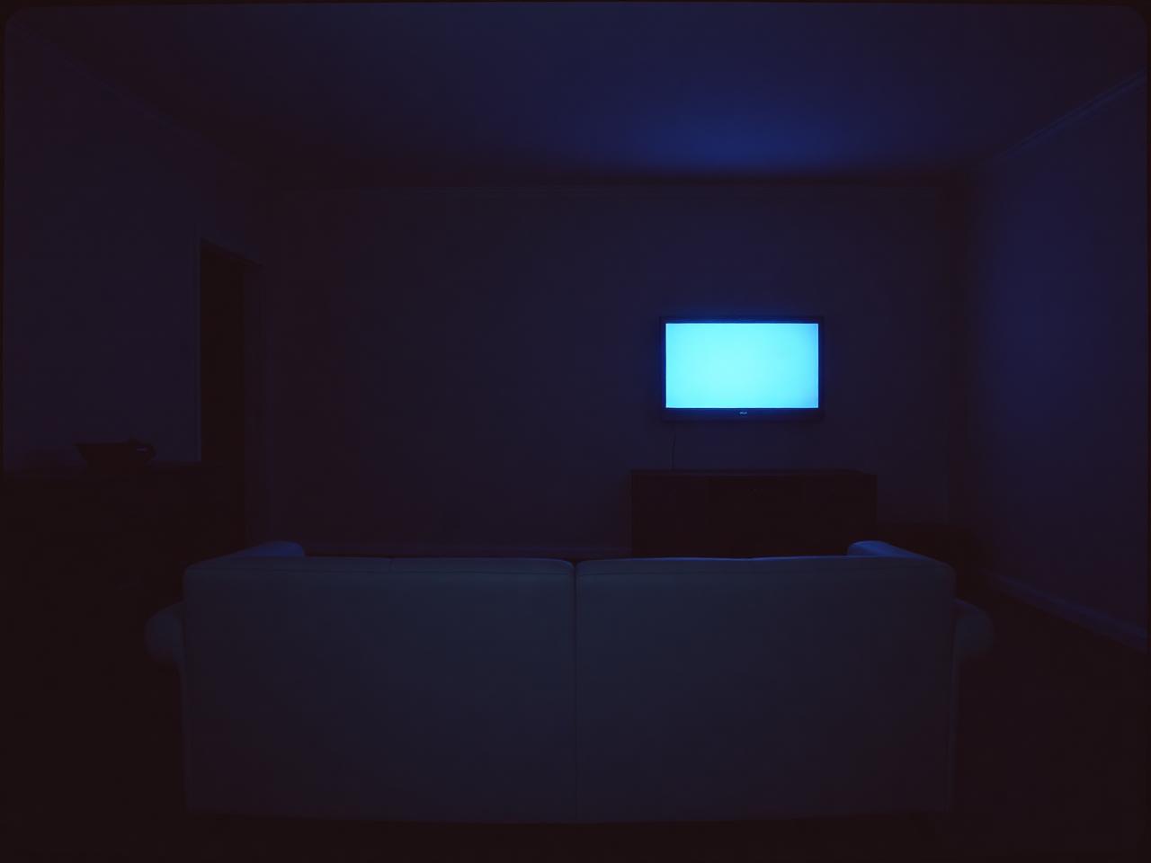 An empty sofa in a dark living room, lit only by the pale flicker of an off-screen television reflected on the far wall.